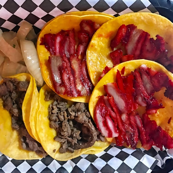 A basket of Tacos El Gordo's tacos, featuring red marinated pork and beef, with pickled onions.